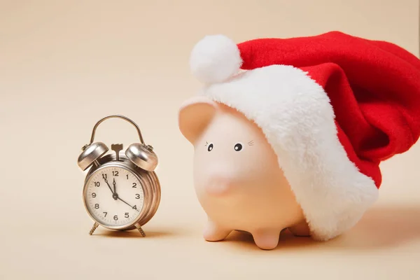 Pink piggy money bank with Christmas hat, alarm clock isolated on pastel beige background. Money accumulation, investment, banking or business services, wealth concept. Copy space advertising mock up