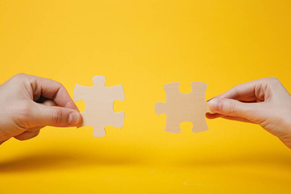 Close up cropped photo of hands holding trying to connect couple wooden jigsaw puzzle pieces isolated on bright yellow wall background. Association, connection concept. Copy space advertising mock up
