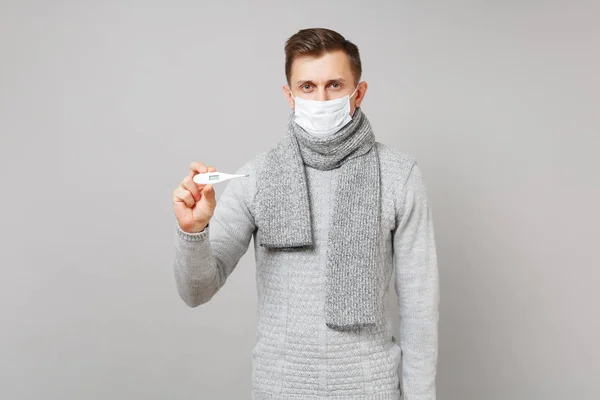 Young man in gray sweater, scarf with sterile face mask holding thermometer isolated on grey background in studio. Healthy lifestyle ill sick disease treatment cold season concept. Mock up copy space