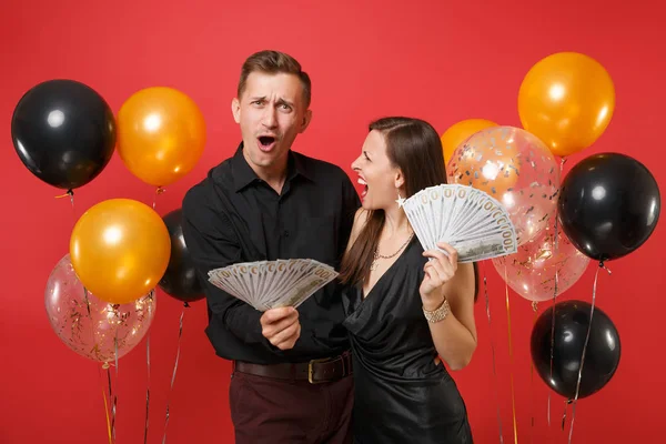 Couple in black clothes celebrating birthday holiday party, hold cash money isolated on bright red background air balloons. St. Valentine International Women Day Happy New Year 2019 concept. Mock up