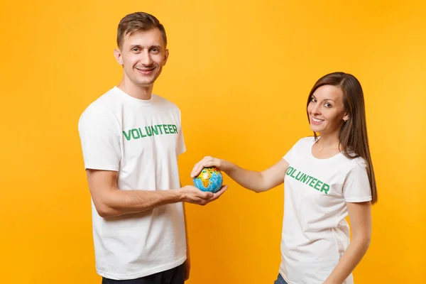 Colleagues couple in white t-shirt with inscription green title volunteer in palms Earth world globe isolated on yellow background. Voluntary free work, assistance help charity grace teamwork concept