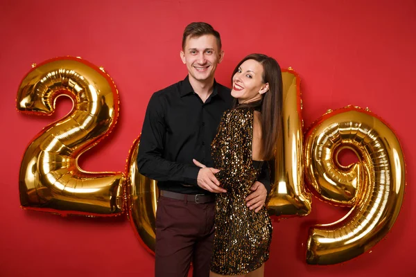 Couple guy girl in shiny glitter dress, black shirt celebrating holiday party isolated on bright red wall background golden numbers air balloons studio portrait. Happy New Year 2019 Christmas concept