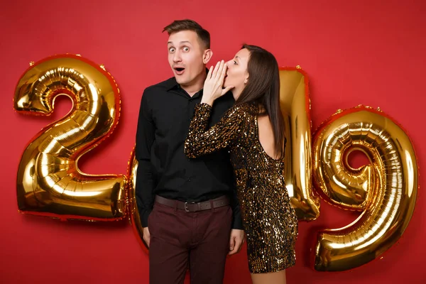 Couple guy girl in shiny glitter dress, black shirt celebrating holiday party isolated on bright red wall background golden numbers air balloons studio portrait. Happy New Year 2019 Christmas concept