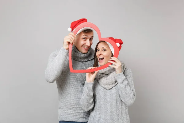 Merry fun couple girl guy in red Santa Christmas hat gray sweaters scarves isolated on grey wall background, studio portrait. Happy New Year 2019 celebration holiday party concept. Mock up copy space