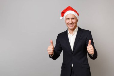 Neşeli genç iş adamı klasik siyah elbiseli, başparmak gösterilmesini Noel şapka gri arka plan üzerinde izole. Başarı kariyer servet iş. Mutlu yeni yıl 2019 kutlama tatil parti konsept
