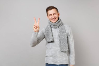 Gri kazak, zafer işareti gösterilen eşarp neşeli genç adam gri arka plan üzerinde stüdyo portre izole. Sağlıklı moda yaşam tarzı, insanlar samimi duygular, soğuk sezon kavramı. Kopyalama alanı alay