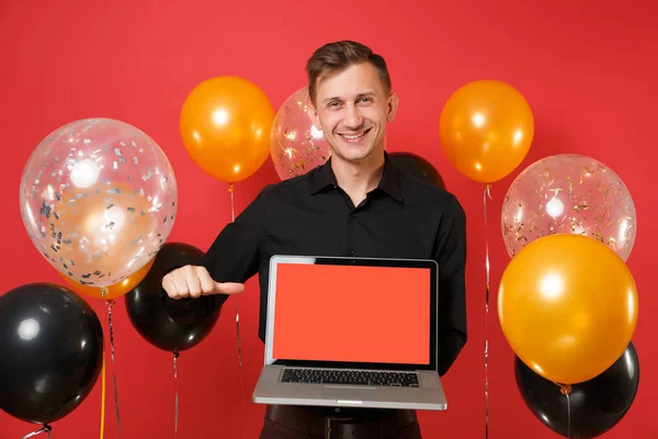 Smiling young man in black classic shirt pointing thumb aside on laptop pc computer with blank black empty screen on red background air balloons. Happy New Year, birthday mockup holiday party concept