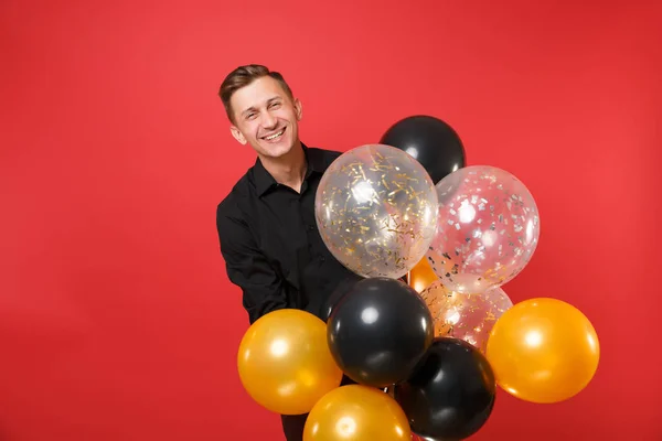 Funny young man in black classic shirt holding air balloons, celebrating isolated on bright red background. Valentine\'s, International Women\'s Day Happy New Year birthday mockup holiday party concept