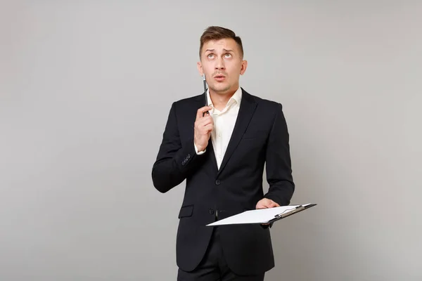 Puzzled young business man in classic black suit looking up, holding pencil, clipboard with papers document isolated on grey background. Achievement career wealth business concept. Mock up copy space