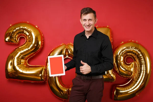Merry fun young man in black shirt celebrating holiday party hold tablet pc isolated on bright red wall background, golden numbers air balloons studio portrait. Happy New Year 2019 Christmas concept