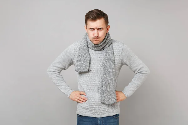 Strenger junger Mann im grauen Pullover, Schal stehend mit den Armen akimbo auf Taille isoliert auf grauem Wandhintergrund. gesunde Mode Lebensstil Menschen aufrichtige Emotionen kalte Jahreszeit Konzept. Attrappe Kopierraum — Stockbild Strenger Junger Mann Grauen Pullover Schal Stehend Mit Den Armen — Stockfoto