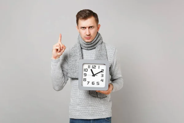 グレーのセーターに若い男は 灰色の背景に分離された正方形の時計を持って 人差し指スカーフします 健全なライフ スタイル の心から感情 寒い季節の概念 コピー スペースをモックアップします — ストック写真