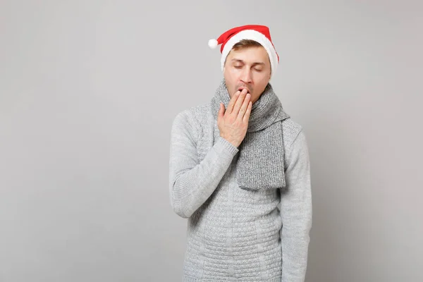 Sleepy Santa man in gray sweater scarf Christmas hat yawning, covering mouth with hand isolated on grey background in studio. Happy New Year 2019 celebration holiday party concept. Mock up copy space