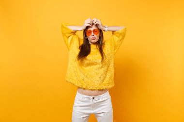 Kürk kazak kalp turuncu gözlük aptal dudakları parlak sarı arka plan üzerinde izole kafasına ellerini koyarak üfleme etrafında komik genç kadın. İnsanlar samimi duygular, yaşam kavramı. Reklam alanı