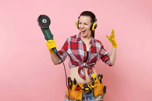 Strong energy handyman woman in yellow gloves, noise insulated headphones, kit tools belt full of instruments holding power saw isolated on pink background. Female in male work. Renovation concept