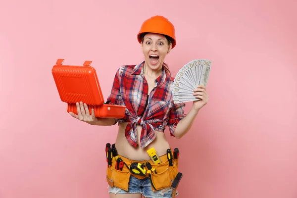 Strong woman in orange helmet, plaid shirt, denim shorts, kit tools belt full of instruments, toolbox, lots cash money isolated on pink background. Female in male work. Renovation occupation concept