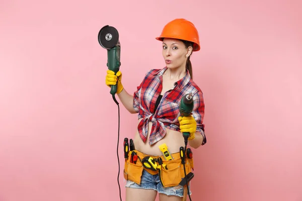 Energy fun handyman woman in gloves, protective orange helmet, kit tools belt full of instruments holding power saw electric drill isolated on pink background. Female in male work. Renovation concept