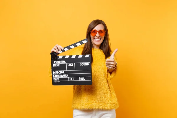 Smiling young woman in orange heart eyeglasses showing thumb up, holding classic black film making clapperboard isolated on yellow background. People sincere emotions, lifestyle. Advertising area
