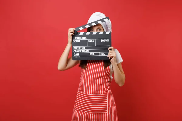 Housewife female chef cook or baker in striped apron, white t-shirt, toque chefs hat isolated on red wall background. Woman holding classic black film making clapperboard. Mock up copy space concept