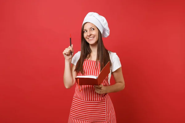 Housewife female chef cook or baker in striped apron white t-shirt toque chefs hat isolated on red wall background. Smiling woman hold notepad recipe cooking book and pen. Mock up copy space concept