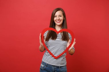 Büyük kırmızı ahşap kalp parlak kırmızı duvar arka plan üzerinde izole tutarak rahat çizgili kıyafetler içinde gülümseyen güzel genç kadın portresi. İnsanlar samimi duygular, yaşam kavramı. Kopyalama alanı alay