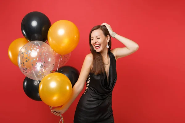 Happy girl with closed eyes in black dress celebrating putting hand on head holding air balloons isolated on red background. St. Valentine\'s Day, Happy New Year, birthday mockup holiday party concept