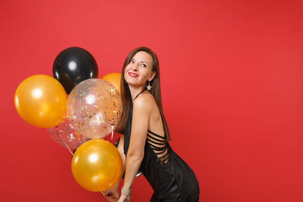 Smiling young woman in little black dress celebrating looking up and holding air balloons isolated on bright red background. St. Valentine's Day, Happy New Year, birthday mockup holiday party concept