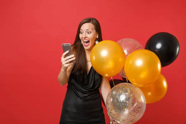 Excited happy young girl in little black dress holding air balloons using mobile phone while celebrating isolated on red background. Women\'s Day, Happy New Year, birthday mockup holiday party concept