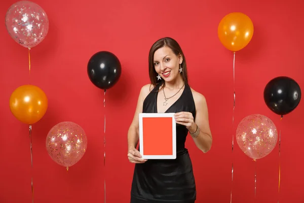Beautiful young woman in black dress celebrating holding tablet pc computer with blank black empty screen on bright red background air balloons. Happy New Year, birthday mockup holiday party concept