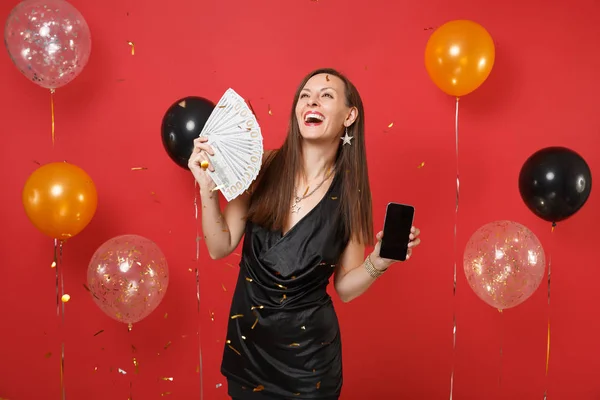 Laughing young woman holding mobile phone with blank empty black screen bundle lots of dollars, cash money on bright red background air balloons. Happy New Year, birthday mockup holiday party concept