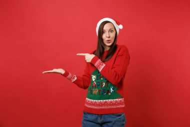 Örme kazak hayran Noel Baba kız, Noel şapka seyir işaret eden bir ele, bir kenara izole kırmızı zemin üzerine parmak şaşırttı. Mutlu yeni yıl 2019 kutlama tatil parti kavramı. Kopyalama alanı alay