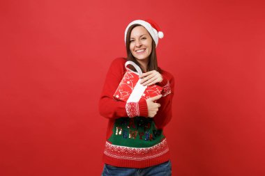 Örme kazak genç Noel Baba kız gülümseyerek, Noel şapka hediye kırmızı kutu ile sarılma üzerinde izole kırmızı arka plan mevcut. Mutlu yeni yıl 2019 kutlama tatil parti kavramı. Kopyalama alanı alay