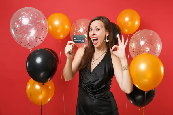 Excited young woman in little black dress celebrating, holding credit card, showing OK sign on red background air balloons. St. Valentine\'s Day, Happy New Year, birthday mockup holiday party concept