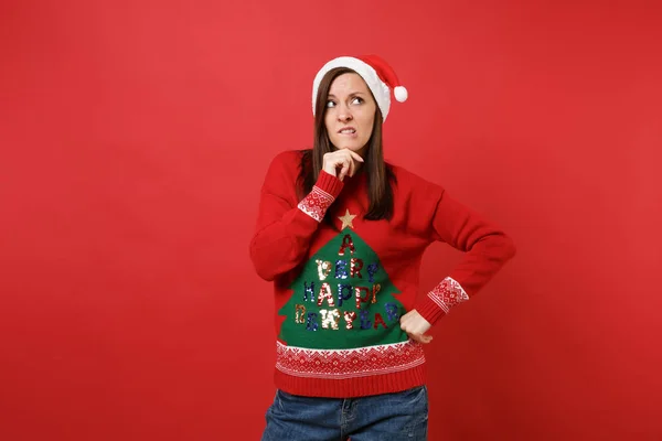 Puzzled pensive young Santa girl in Christmas hat looking up put hand prop up on chin biting lips isolated on red background. Happy New Year 2019 celebration holiday party concept. Mock up copy space