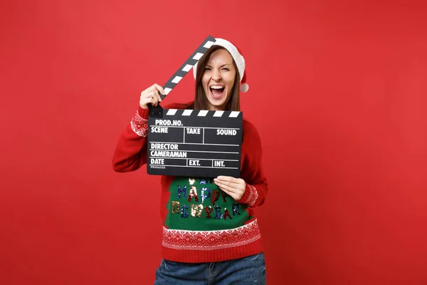 Cheerful young Santa girl in Christmas hat screaming, hold classic black film making clapperboard isolated on red background. Happy New Year 2019 celebration holiday party concept. Mock up copy space