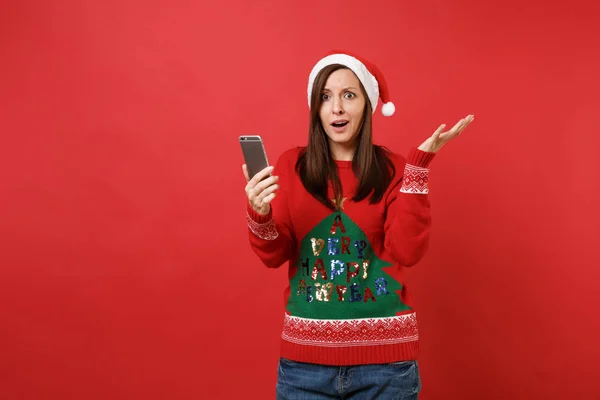 ニットのセーター クリスマス帽子の両手赤の背景に分離した携帯電話の普及でサンタ少女の困惑 幸せな新しい年 2019年お祝いの休日党の概念 コピー スペースをモックアップします — ストック写真