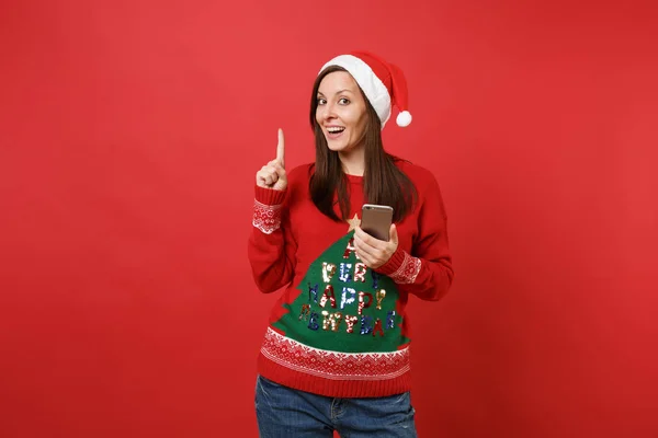 クリスマス帽子人差し指を携帯電話を保持しているセーターのサンタ少女の笑みを浮かべて 赤の背景に分離されました 幸せな新しい年 2019年お祝いの休日党の概念 コピー スペースをモックアップします — ストック写真