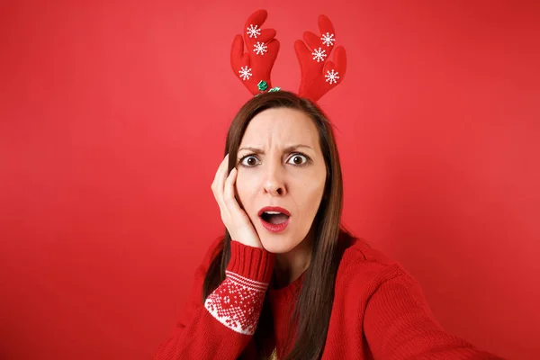Close up selfie shot of puzzled young Santa girl in fun decorative deer horns put hand on cheek isolated on red background. Happy New Year 2019 celebration holiday party concept. Mock up copy space