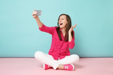 Gül gömlek, beyaz pantolon selfie cep telefonundan atış yapıyor katta oturan kadında tam uzunlukta parlak pembe mavi pastel duvar arka plan studio izole. Moda yaşam tarzı kavramı. Kopyalama alanı alay