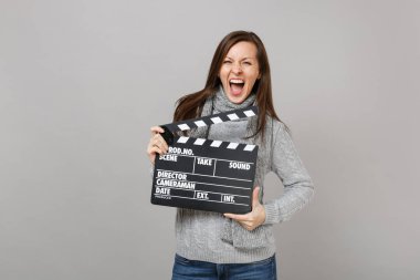 Crazy young woman in gray sweater, scarf screaming, holding classic black film making clapperboard isolated on grey background. Healthy fashion lifestyle, people sincere emotions, cold season concept clipart
