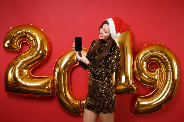 Merry Santa girl in shiny glitter dress, Christmas hat hold mobile phone isolated on bright red wall background, golden numbers air balloons studio portrait. Happy New Year 2019 holiday party concept