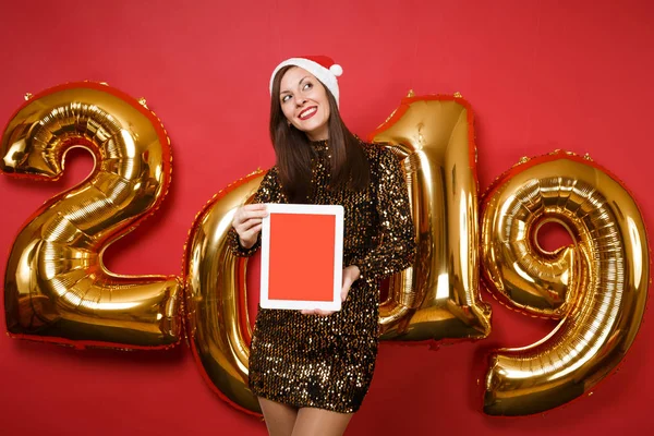 Merry Santa girl in shiny glitter dress, Christmas hat hold tablet pc isolated on bright red wall background, golden numbers air balloons studio portrait. Happy New Year 2019 holiday party concept