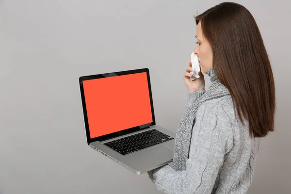 Side view of woman in sweater scarf with paper napkin holding laptop pc computer with blank empty screen isolated on grey background. Healthy lifestyle online treatment consulting cold season concept