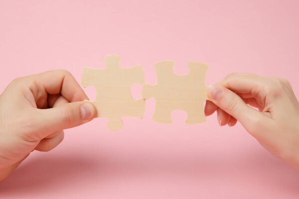 Close up cropped photo of hands holding trying to connect couple wooden jigsaw puzzle pieces isolated on pastel pink wall background. Association, connection concept. Copy space advertising mock up