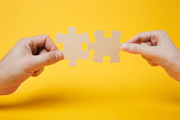 Close up cropped photo of hands holding trying to connect couple wooden jigsaw puzzle pieces isolated on bright yellow wall background. Association, connection concept. Copy space advertising mock up