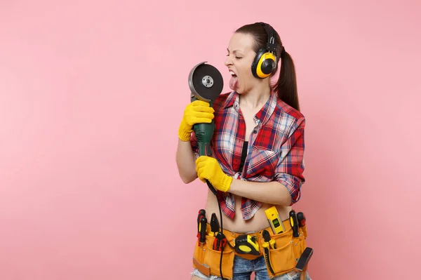 Strong energy handyman woman in yellow gloves, noise insulated headphones, kit tools belt full of instruments holding power saw isolated on pink background. Female in male work. Renovation concept