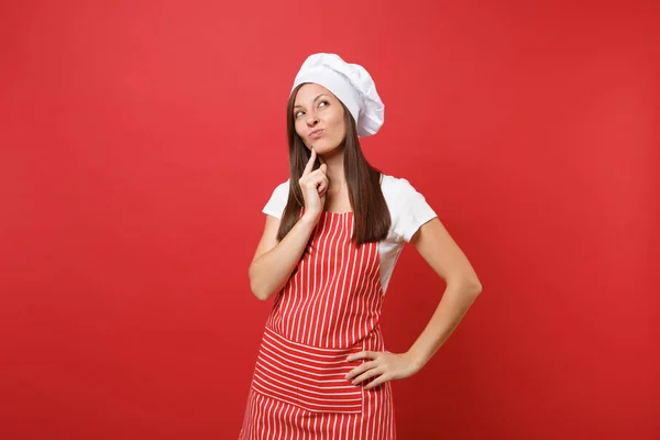 Housewife female chef cook or baker in striped apron, white t-shirt, toque chefs hat isolated on red wall background. Beautiful housekeeper woman put hand prop up on chin. Mock up copy space concept