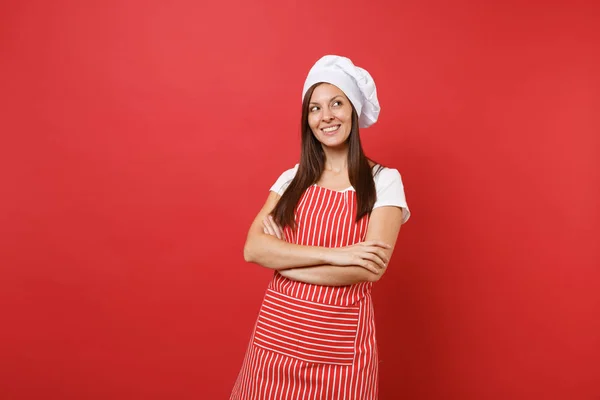 Housewife female chef cook or baker in striped apron, white t-shirt, toque chefs hat isolated on red wall background. Pretty young housekeeper woman holding hands crossed. Mock up copy space concept