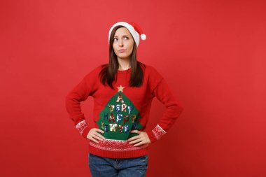 Santa kız ayakta izole kırmızı zemin üzerine silah akimbo ile bakılması örme kazak Noel şapkalı şaşkın. Mutlu yeni yıl 2019 kutlama tatil parti kavramı. Kopyalama alanı alay