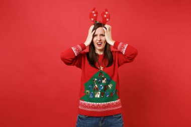 Örme kazak içinde memnun Noel Baba kız ellerini kafasına izole kırmızı zemin üzerine koyarak dekoratif geyik boynuzları eğlenceli. Mutlu yeni yıl 2019 kutlama tatil parti kavramı. Kopyalama alanı alay